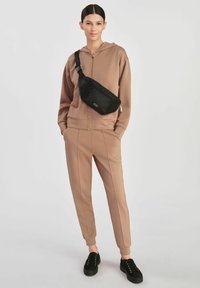 Miękka, brązowa bluza zapinana na zamek i dopasowane, zwężane dresowe spodnie. Czarna nerka z teksturowanym wzorem noszona na ciele. Czarne sneakersy.