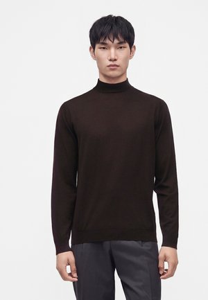 Junger Mann mit kurzen schwarzen Haaren, der einen eng anliegenden schwarzen Rollkragenpullover und dunkelgraue Hosen trägt, steht vor einem schlichten weißen Hintergrund.