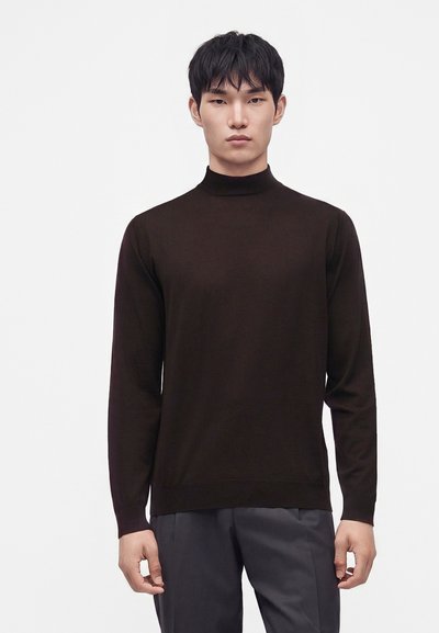 Jeune homme aux cheveux courts et noirs portant un pull noir à col roulé ajusté et un pantalon gris foncé, debout devant un fond blanc uni.