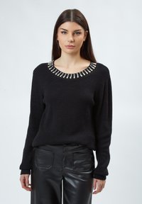 Zwarte gebreide trui met een wijde halslijn, versierd met kraalaccenten, gedragen met zwarte nepleer broek. Gelaagd materiaal en een relaxte pasvorm.