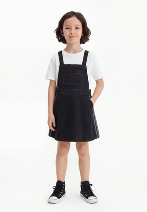 Bambino con capelli scuri fino alle spalle, che indossa una maglietta bianca, un vestito salopette in denim nero e sneakers alte nere, in piedi con una mano in tasca.