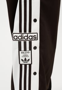 Nahaufnahme von schwarzen Adidas-Trainingshosen mit weißen Seitenstreifen, Druckknöpfen und Aufnähern mit den Texten "adidas" und "The Brand with the Three Stripes".