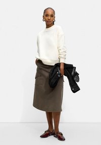 Kvinne iført en hvit genser, olivengrønn knelang skjørt, burgunder leiligheter, og holder en svart jakke mot en ensfarget bakgrunn.