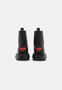 Sorte ankelboots i svart lær med en kraftig såle, med detalj av rød logo bak og stropper for enkel påføring. Glatt tekstur.
