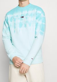 Jasnoniebieska bluza tie-dye wykonana z bawełny, z prążkowanymi mankietami i dekoltem. Posiada wyszywane logo "TOMMY JEANS" oraz mały akcent z logo na rękawie.