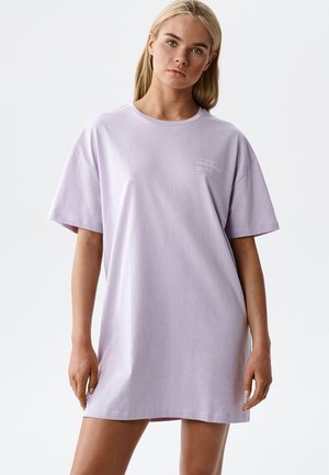 Blondynka w luźnej, jasnofioletowej, oversize'owej sukience w stylu T-shirt, stojąca na tle jednolitej białej ściany.