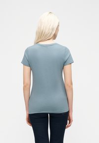 Γυναίκα με μακριά ξανθά μαλλιά που φοράει απλό ανοιχτό μπλε κοντομάνικο μπλουζάκι και σκούρο τζιν, όρθια με την πλάτη προς το φακό, μπροστά σε λευκό φόντο.