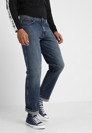Personne portant un jean bleu foncé retroussé, un t-shirt noir à manches longues avec manches imprimées, et des baskets montantes bleu marine, debout.