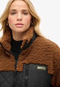 Veste en polaire marron avec tissu matelassé noir ; présente un col montant, des poches zippées et un écusson logo sur le devant. Design texturé et chaud.