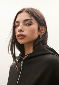 Felpa nera con zip e coulisse, caratterizzata da una texture liscia e cappuccio. Primo piano di una persona con capelli castani di media lunghezza e orecchini.