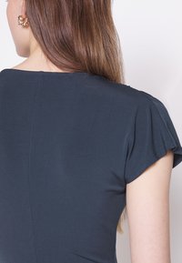 Mörkblå, figursydd topp med en slät struktur, korta, lätta ärmar och en subtil halsringning. Har en detalj med söm bak.