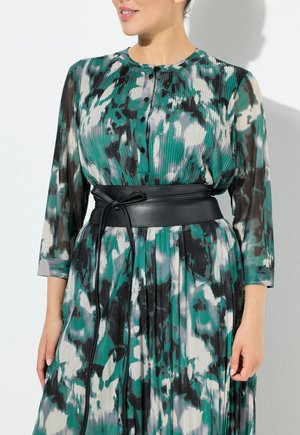 Una cintura nera in pelle stringe un abito lungo e fluido con un motivo nei toni di verde, grigio e bianco, dotato di maniche lunghe plissettate e chiusura a bottoni.