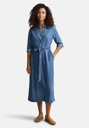 Vrouw met krullend haar die een lange blauwe denim jurk met knopen draagt, met opgerolde mouwen, een ceintuur in de taille en bruine platte schoenen, rechtopstaand.