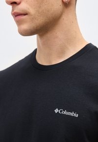 Κοντινή λήψη ενός ατόμου που φοράει μαύρο t-shirt Columbia με μικρό λευκό λογότυπο στο στήθος και ασημί κρίκο στο αριστερό αυτί.