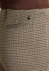 Bikses brūnā un bēšā pundurzvēra rakstā ar teksturētu audumu. Tās ir ar aizmugures kabatu un melnu izšūtu detaļu virs kabatas.