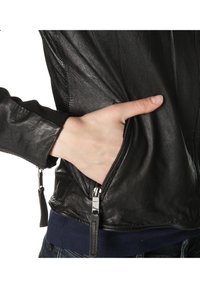 Main insérée dans la poche zippée d'une veste en cuir noire portée sur une chemise bleu foncé et un jean.