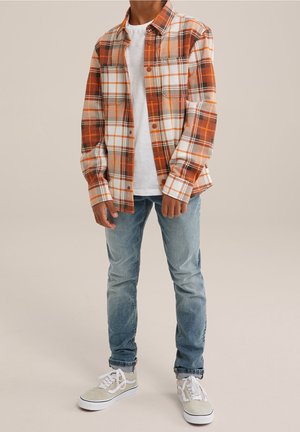 Person trägt orange-weiß kariertes Hemd mit Knöpfen, weißes T-Shirt, hellblaue Jeans und beige Turnschuhe, steht vor einfachem Hintergrund.