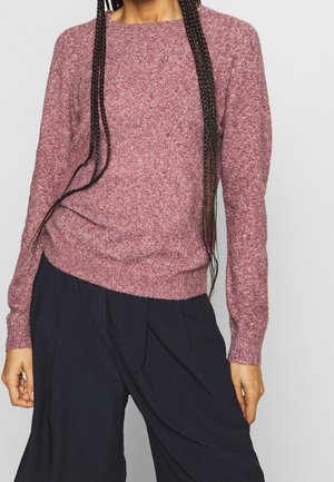 Personne portant un pull en maille mauve à manches longues et un pantalon noir ample, avec de longs cheveux tressés tombant devant.