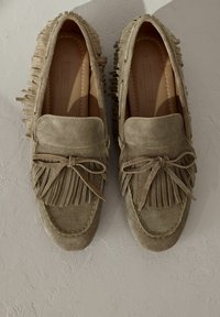 Mocasines de ante en beige claro con detalles de flecos y acento de lazo. Presentan una punta redonda y una suela cosida para añadir textura.