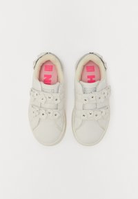 Zapatillas de cuero blanco con acentos en forma de flor, correas de velcro y una plantilla rosa. Presentan una punta redondeada y detalles metálicos sutiles.