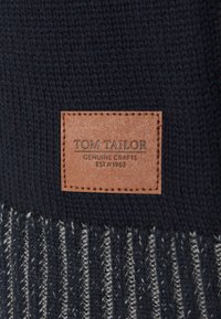 Écharpe tricotée bleu marine avec une étiquette en cuir portant un texte embossé. Le motif comprend des rayures alternées gris clair et bleu marine foncé.