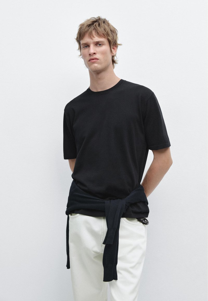 Ung mand med lyst brunt hår iført en sort t-shirt med korte ærmer, hvide bukser og en sort trøje bundet om livet.