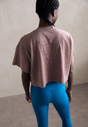 Kvinde med flettet hår iført en løs brun cropped top med et broderet tyr-design og lyseblå leggings, stående mod en ensfarvet baggrund.