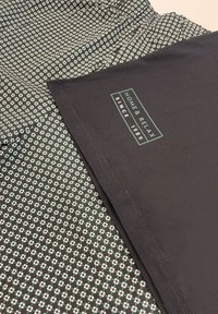 Czarna tkanina z białym i zielonym wzorem geometrycznym, z metką: "DOM I RELAKS OD 1989 R." Miękka tekstura, swobodny design.