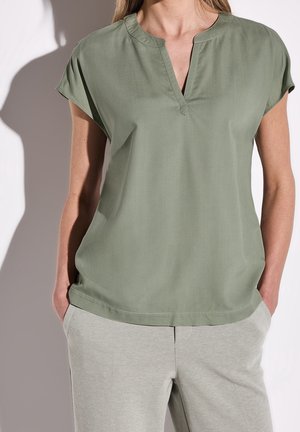 Kobieta ubrana w jasnozieloną bluzkę z krótkim rękawem i rozciętym dekoltem oraz jasnoszare spodnie, ręce w kieszeniach, na jednolitym tle.