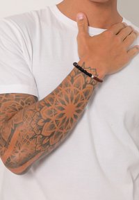 Bracelet en perles noires et brunes porté au poignet, présentant un mélange de textures lisses et mates. Design de tatouage visible sur l'avant-bras.