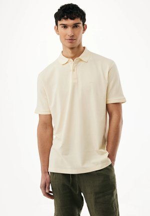 Junger Mann trägt ein schlichtes, cremeweißes Polohemd mit kurzen Ärmeln und dunkelgrüne Hose, steht mit einer Hand in der Tasche.