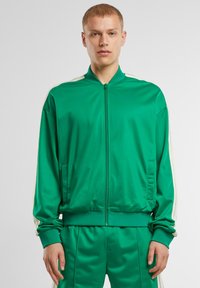 Chaqueta de chándal de satén verde con cuello alto, cierre de cremallera en el frente y detalles en crema en los hombros. Cuenta con bolsillos laterales y puños acanalados.