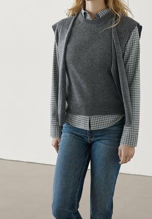 Woman wearing grey sleeveless sweater over black-and-white checkered shirt and blue jeans, standing against a plain light background.