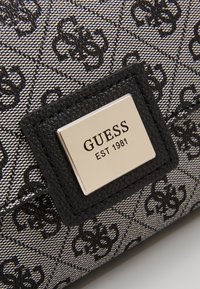 Bolso de mano negro y plateado con una tela sintética estampada, que presenta una notable placa de logo plateado y acentos texturizados en negro.