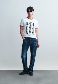 Uomo con occhiali da sole neri, maglietta bianca con stampe di scarpe con tacco alto, jeans blu e scarpe nere, in piedi davanti a uno sfondo uniforme.