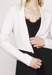 White long-sleeve cardigan with a textured finish and a deep V-neck over a black lace top, paired with black trousers.