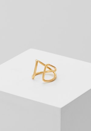 Złoty metalowy pierścionek z otwartym wzorem, na którym znajdują się przecinające się trójkątne i sercowate kontury na białym piedestale.