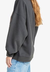 Sudadera oversize gris oscuro con hombros caídos, puños y dobladillo acanalados, que presenta un estampado gráfico en el lateral. Lleva puesta con jeans azules.
