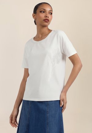 Kvinde iført en hvid kortærmet skjorte med et subtilt blomstermønster og et blåt denimsnit, stående mod en ensfarvet baggrund.