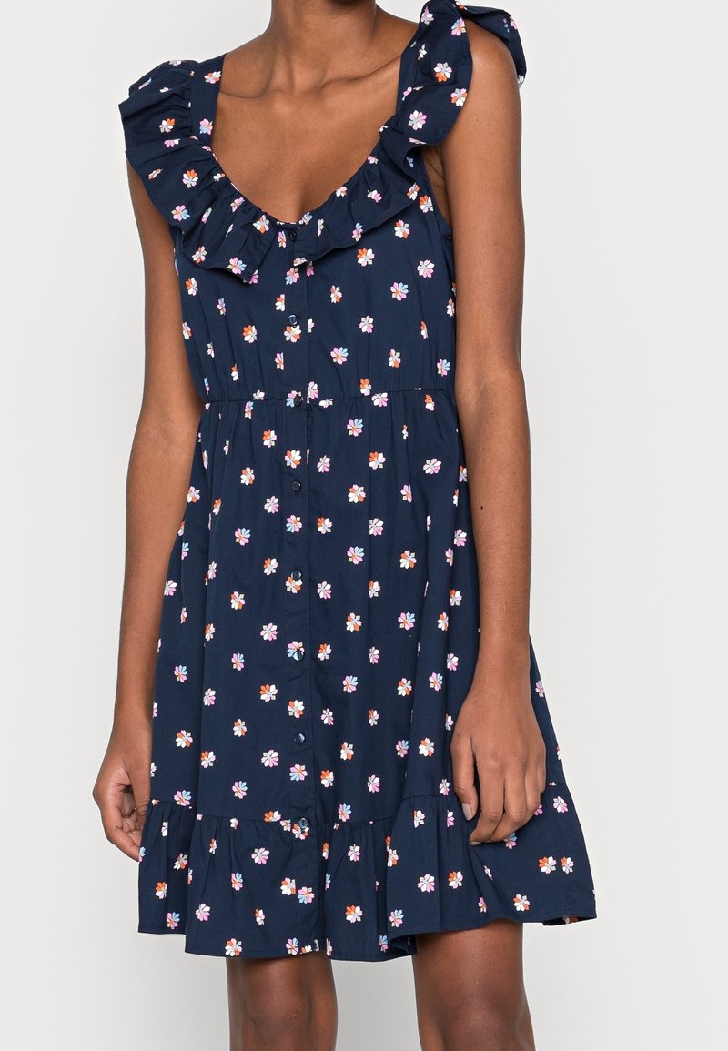 Laivastonsininen kukkamekko, jossa on röyhelöinen pääntie ja olkaimet. Mekossa on pieniä vaaleanpunaisia, oransseja ja valkoisia kukkia. A-linjainen muoto ja kerroksittainen helma.