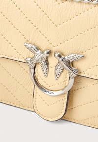 Bolsa de mão de pele amarela acolchoada com ferragens prateadas, apresentando dois acentos de pássaro, um anel circular e detalhes de costura contrastantes.