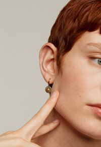 Pendientes de aro dorados con un diseño redondo de estallido solar, con pequeños acentos circulares. Llevados por un modelo, destacando los detalles de la joyería.