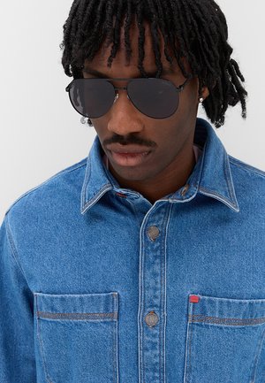 Uomo con capelli neri ricci che indossa occhiali da sole scuri e una giacca di denim blu con tasche sul petto e una piccola etichetta rossa.