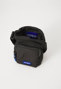 Svart stoffveske med teksturert overflate, glidelåslukking og blå "HUGO"-logo. Har en kompakt form og justerbar stropp.