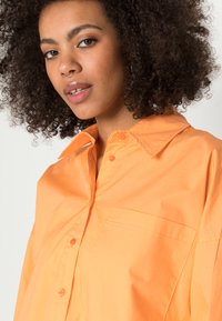 Camisa laranja de botão com colarinho, ajuste oversized e um bolso no peito. O tecido parece suave e leve, com textura mate.