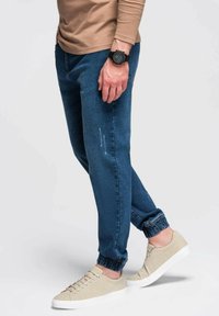 Pantalones jogger de mezclilla azul con puños elásticos, con un patrón desgastado, combinados con zapatillas beige, vistos de lado.