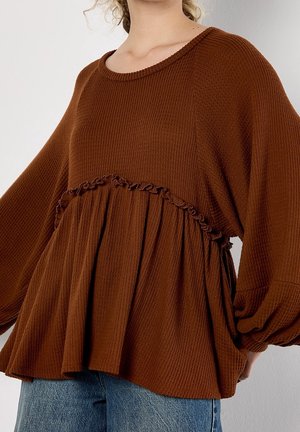 Kvinde iført løs brun tekstureret sweater med flæsedetalje over brystet og ballonærmer, kombineret med blå jeans.