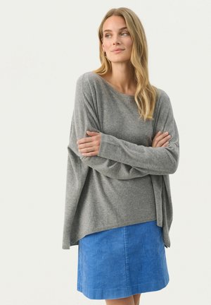 Šedý přes oversized svetr z měkkého materiálu s širokým výstřihem, v kombinaci s modrou kordurovnou sukní. Svetr má dlouhé, volné rukávy.