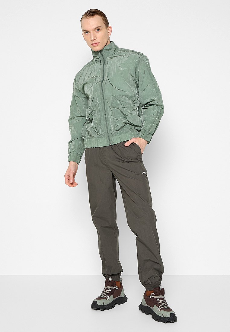 Hellgrüne Bomberjacke mit einem Reißverschluss an der Vorderseite, elastischen Bündchen und zwei Vordertaschen, kombiniert mit dunklen Olivenhosen und robusten braunen Wanderschuhen.