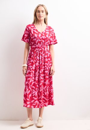 Frau trägt ein rotes und pinkes florales Midikleid mit kurzen Ärmeln und weißen Sneakers, steht vor einem einfarbigen weißen Hintergrund.